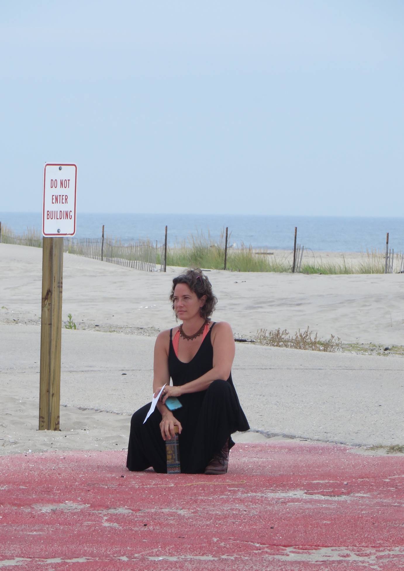 A white woman on a beach facing away from the ocean, crouches in a black sleeveless shirt and skirt wearing a string of black beads wearing a salmon colored scooped neck shirt below. A sign on a wooden 2X4 to her right reads, 